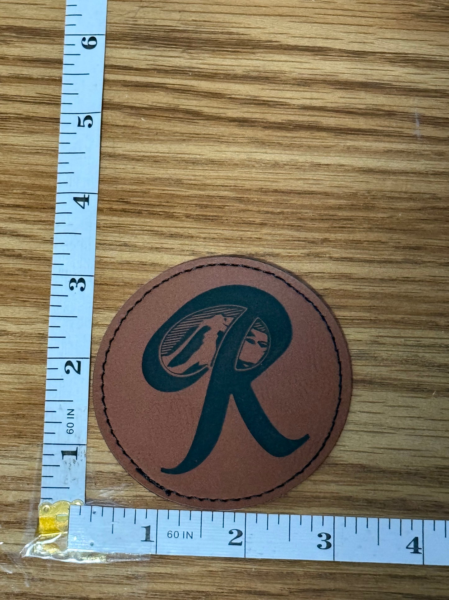 Brown leather patch with a black Rainer logo on a wooden surface with a ruler for scale.