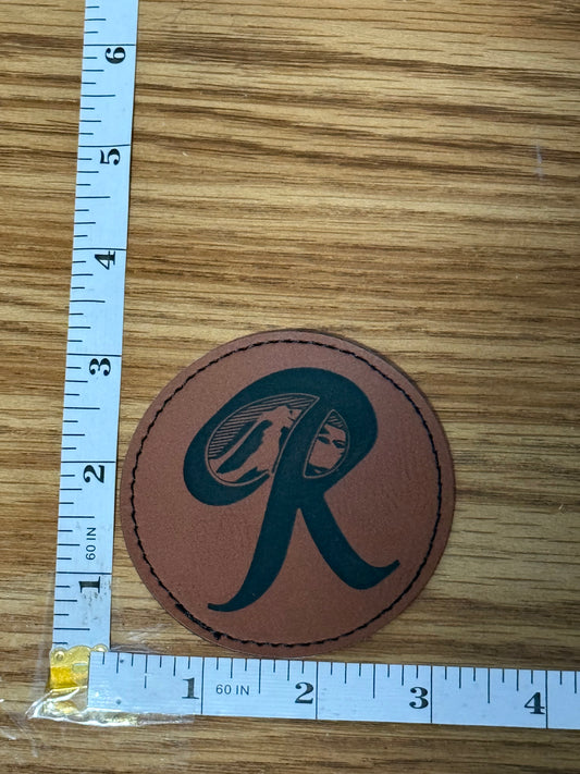 Brown leather patch with a black Rainer logo on a wooden surface with a ruler for scale.