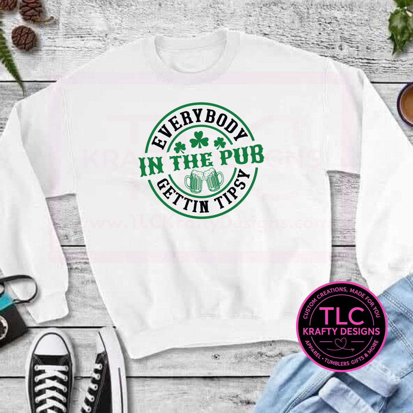 White sweatshirt with green Everybody Gettin' Tipsy Circular Design and shamrock design on a wooden surface, featuring the TLC Krafty Designs logo.