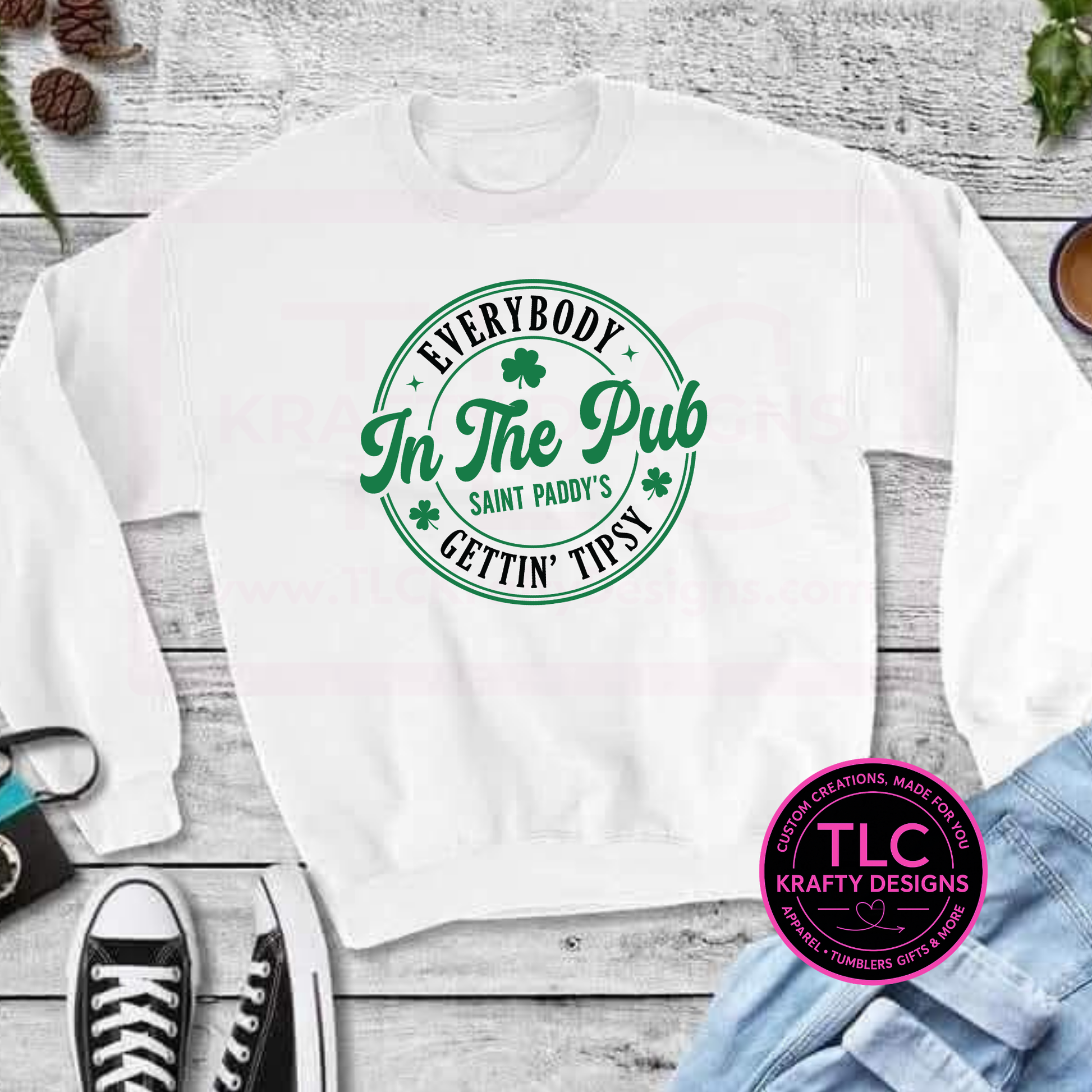 White sweatshirt with green text on a wooden surface, featuring the brand TLC Krafty Designs.